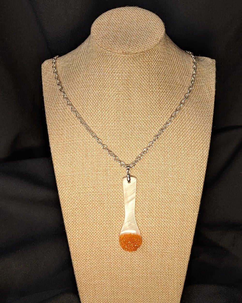 Caviar Spoon Necklace No. 1 — mother-of-pearl spoon with a salmon-orange bowl on a steel chain, on a burlap display bust