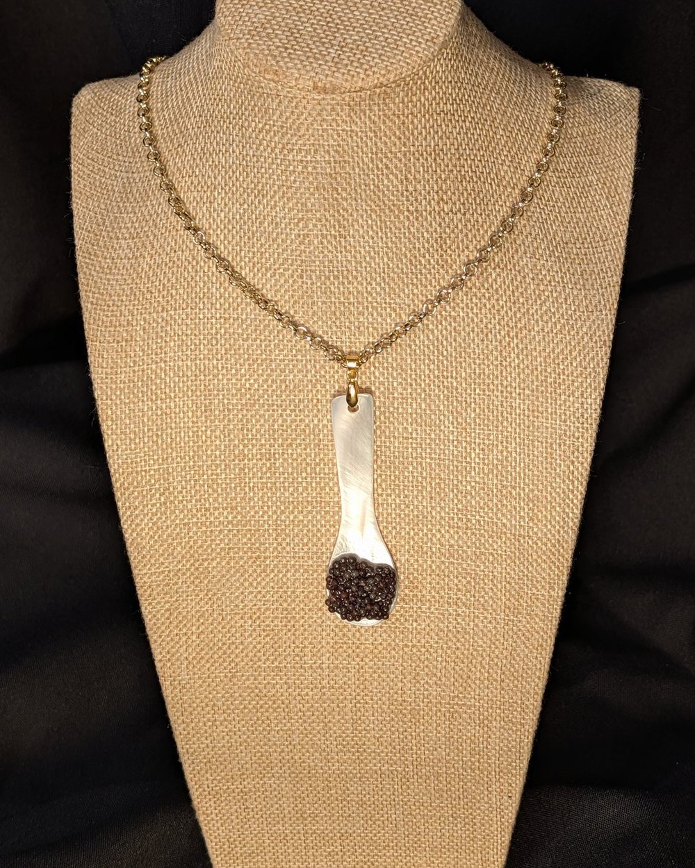 Caviar Spoon Necklace No. 2 — mother-of-pearl spoon with a dark chocolate-brown bowl on a gold-tone chain, on a burlap display bust