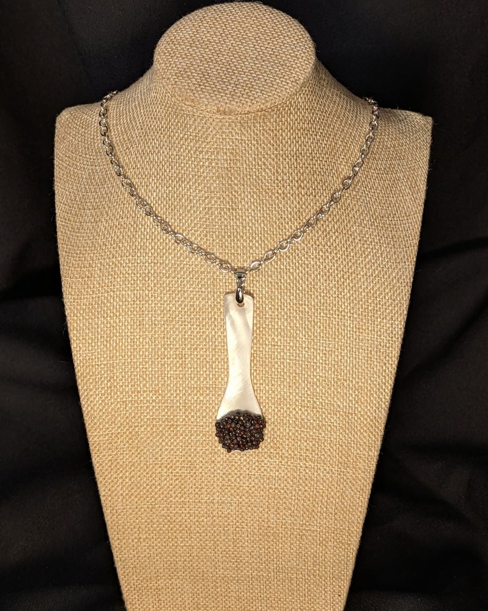 Caviar Spoon Necklace No. 4 — mother-of-pearl spoon with a dark brown bowl on a steel chain, on a burlap display bust