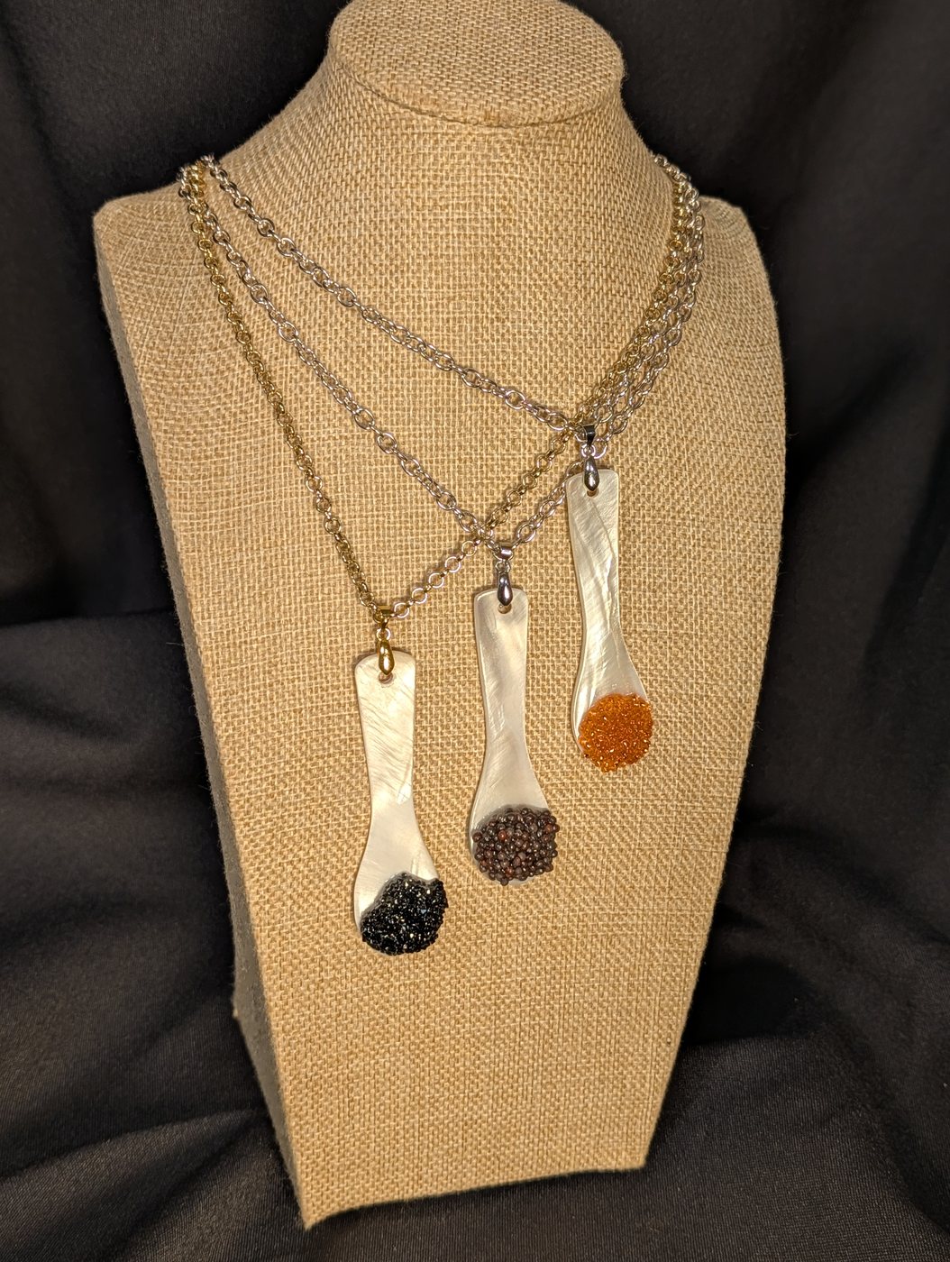 Three mother-of-pearl caviar spoon necklaces on gold and steel chains, hung on a burlap display bust