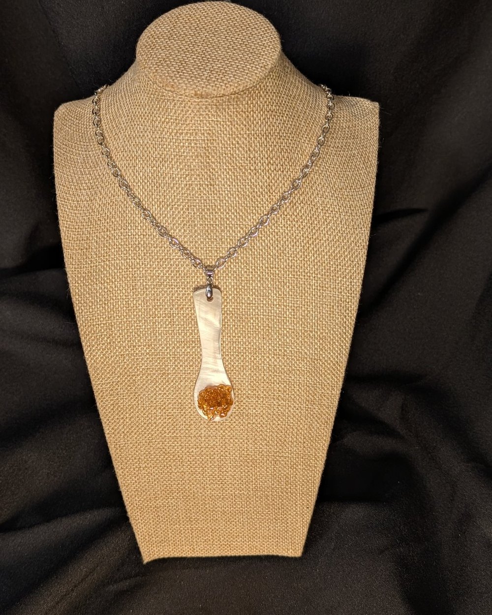Caviar Spoon Necklace No. 5 — mother-of-pearl spoon with a warm amber bowl that catches the light on a steel chain, on a burlap display bust