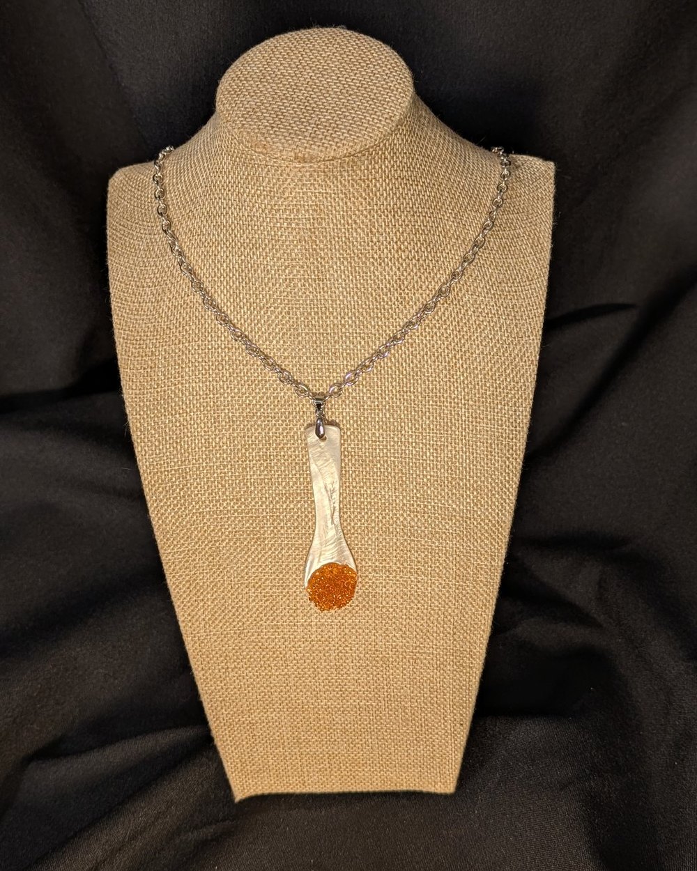Caviar Spoon Necklace No. 6 — mother-of-pearl spoon with a salmon-orange bowl on a steel chain, on a burlap display bust