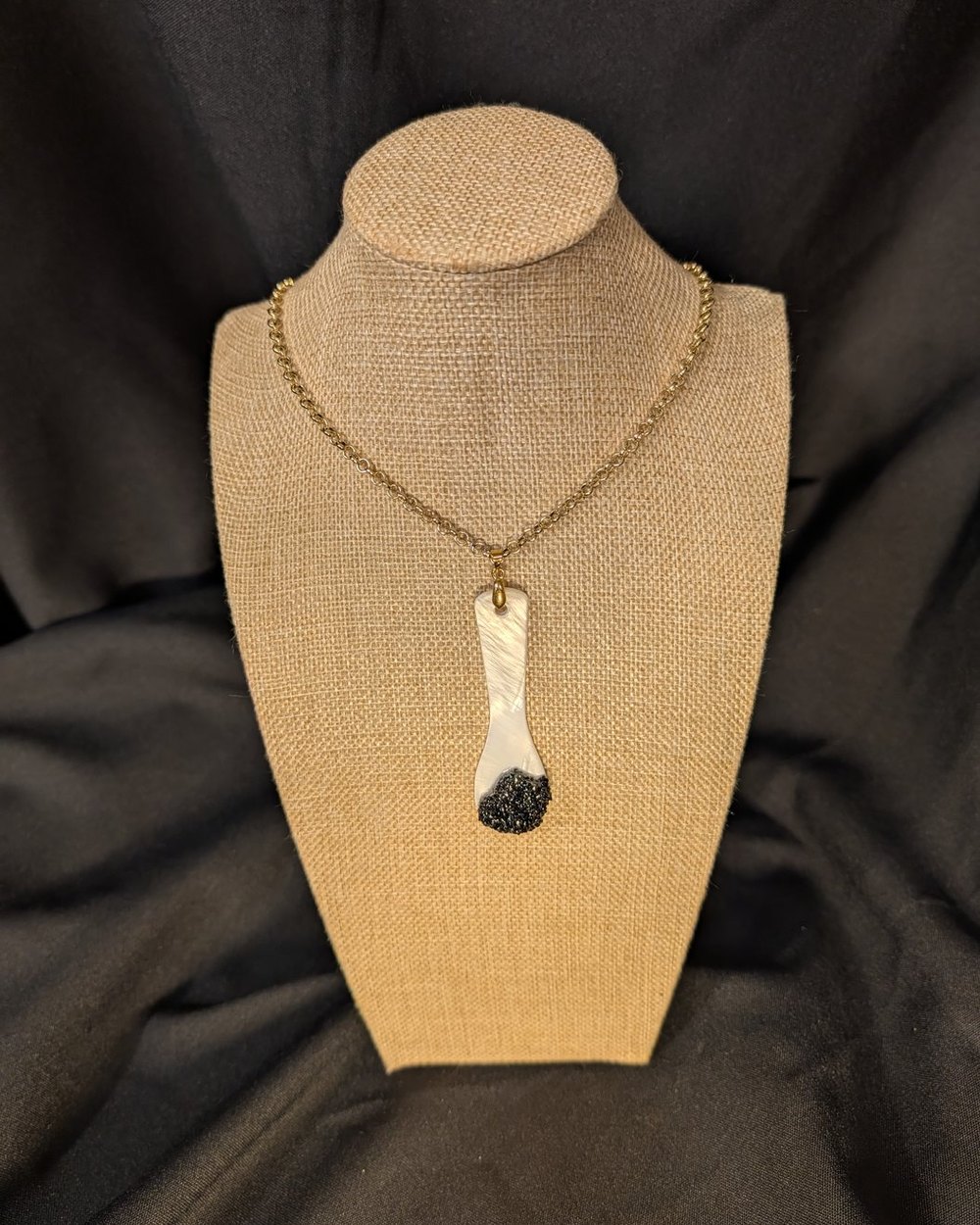 Caviar Spoon Necklace No. 7 — mother-of-pearl spoon with a deep black bowl on a gold-tone chain, on a burlap display bust